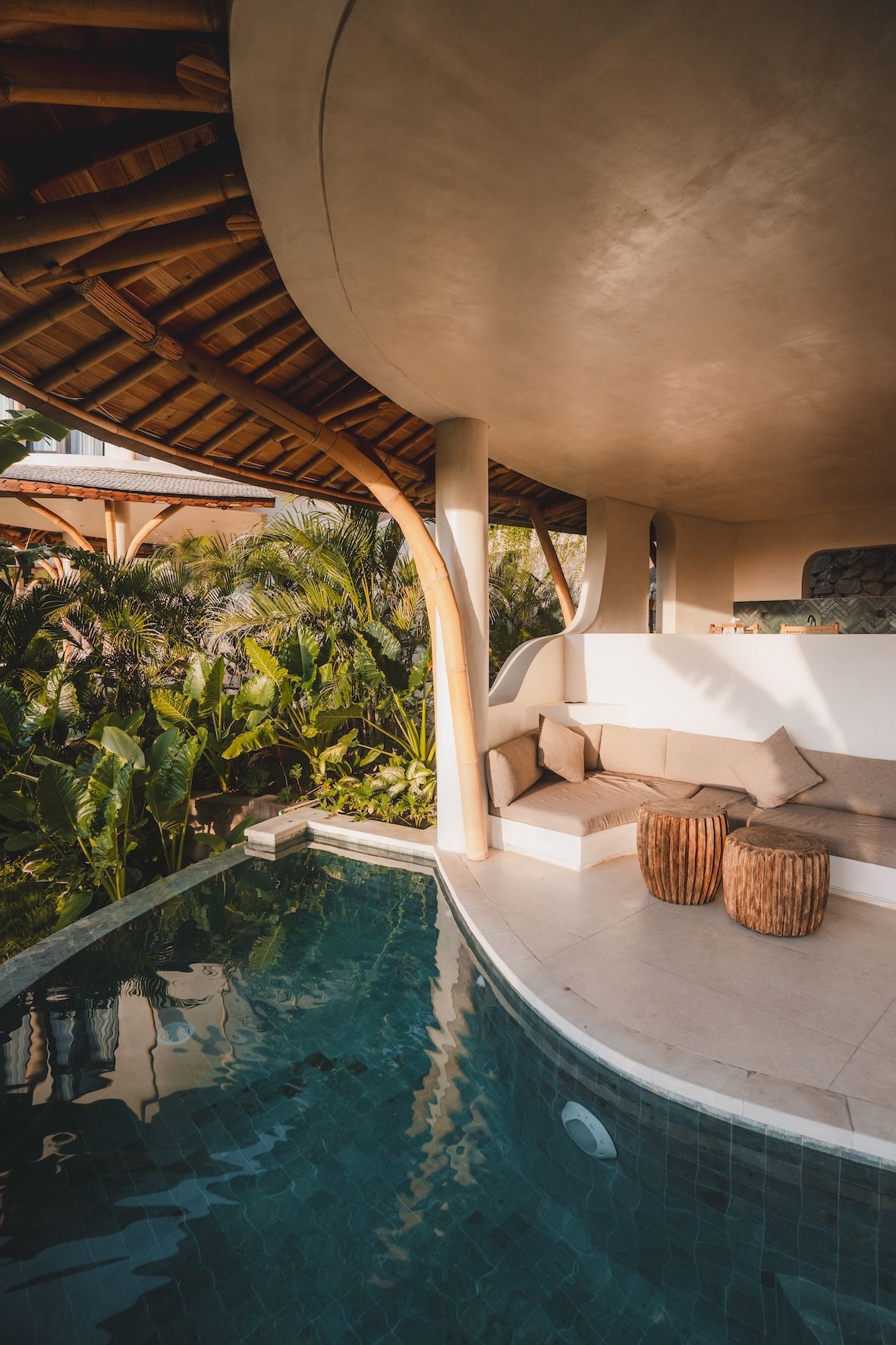 Shaded tropical poolside lounge with curved white sofa, woven tables, and lush greenery beside water
