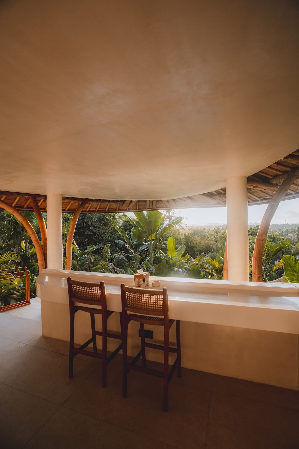 Open-air balcony with a curved ceiling, wooden stools, and a tropical garden view.