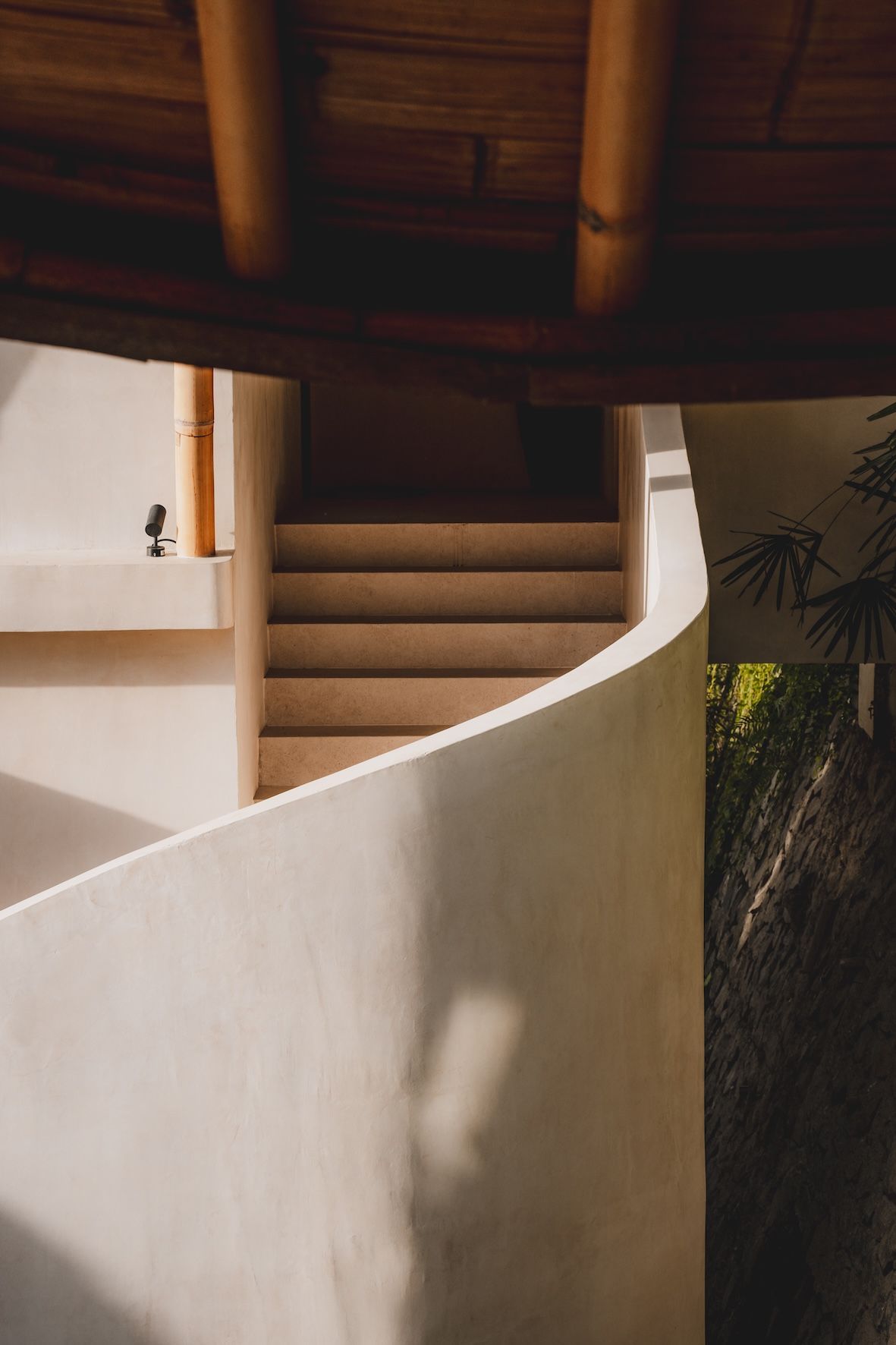 Curved white staircase beneath dark wood beams, with sunlit wall and greenery beside it