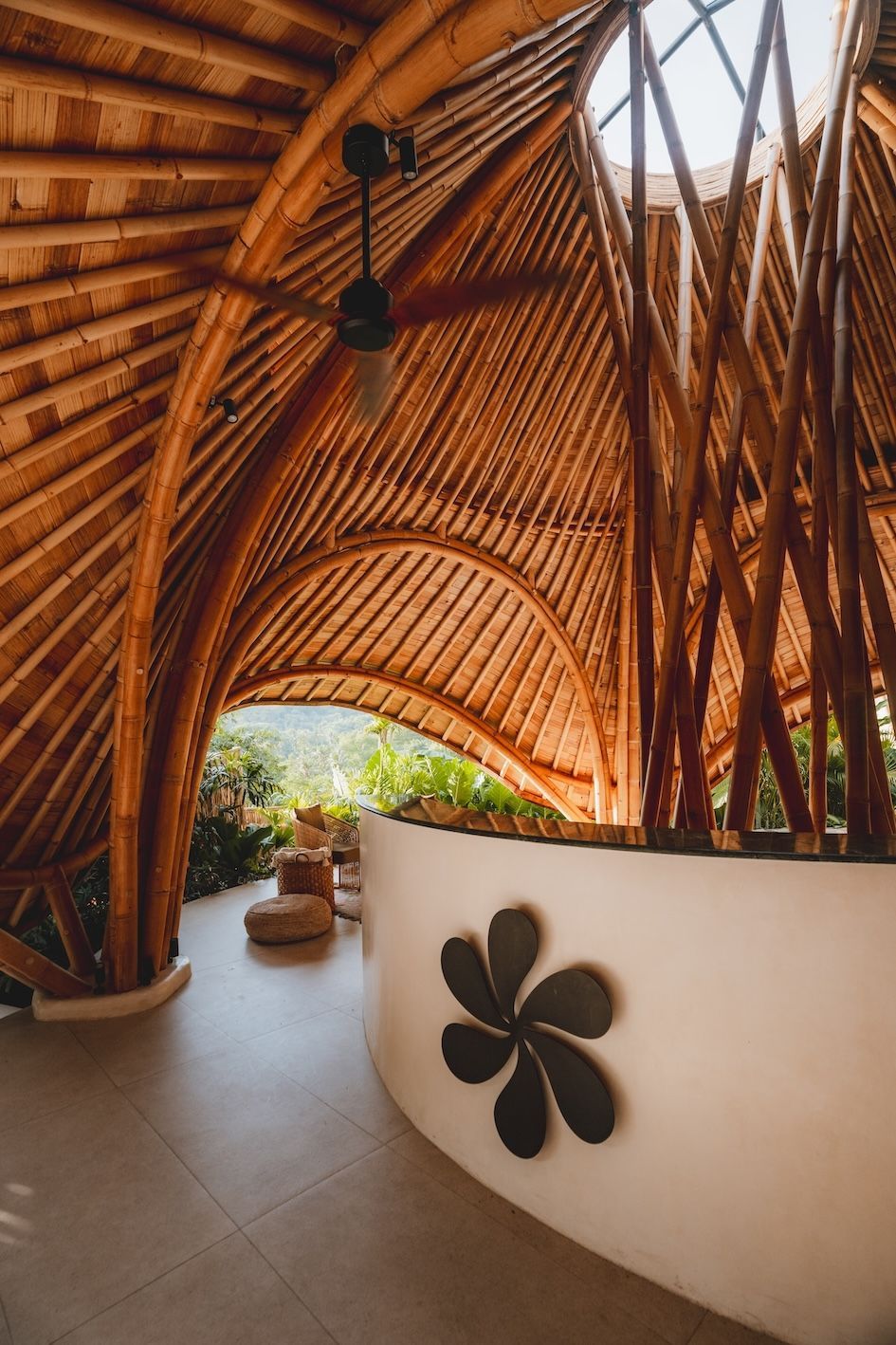 Curved bamboo walkway with arched ceiling, white wall, and potted plants in warm sunlight