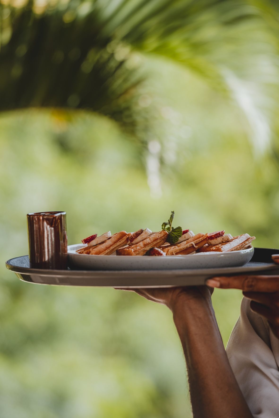 Hand serving a plate of grilled food and a drink on a tray outdoors with blurred greenery behind