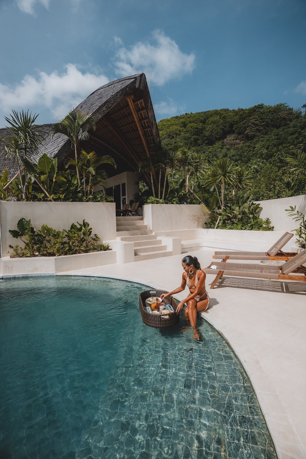Woman sitting by a turquoise pool beside white lounge chairs and tropical greenery