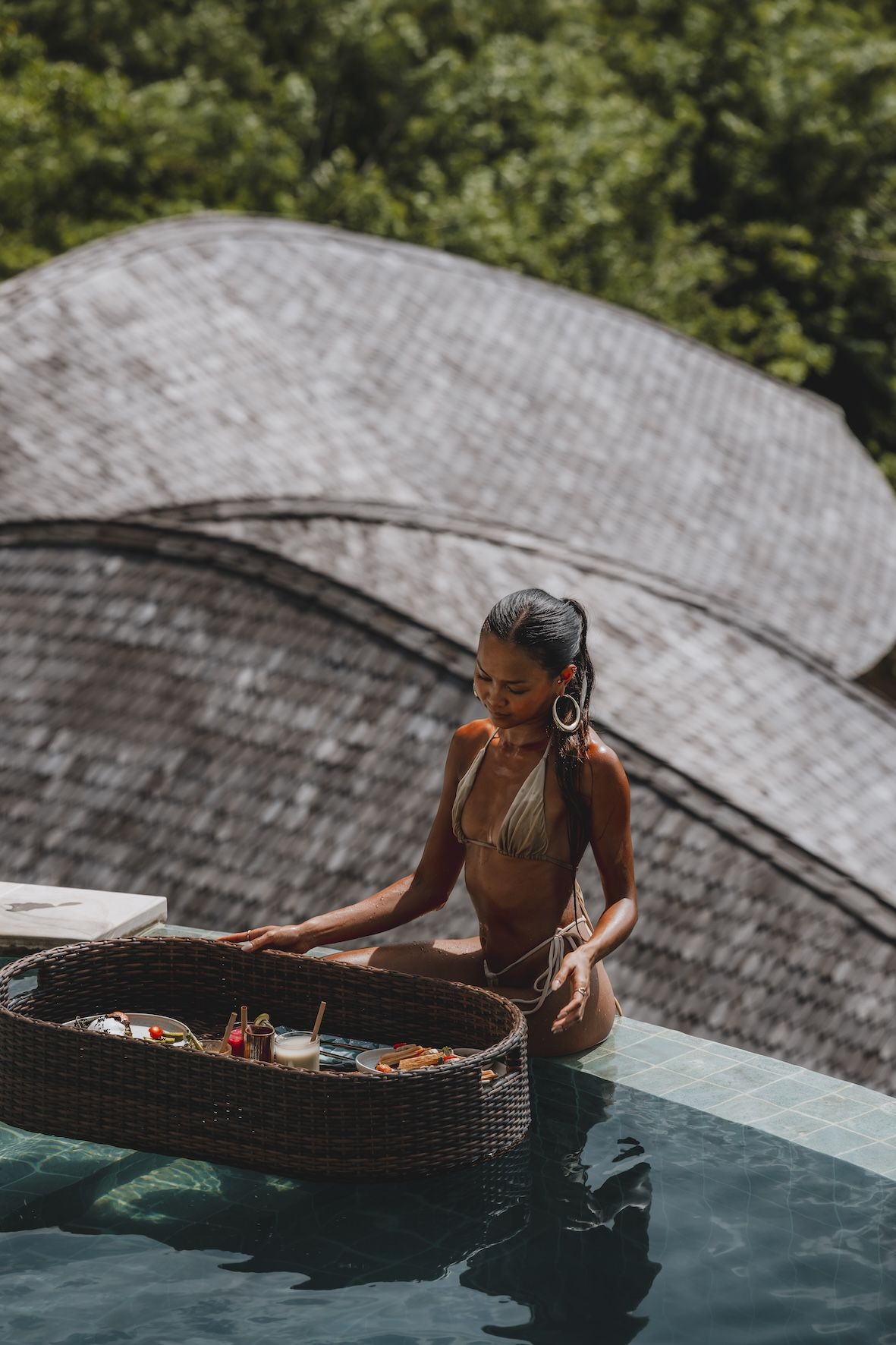 Person standing in a pool with a woven tray, in front of curved gray rooftops and greenery
