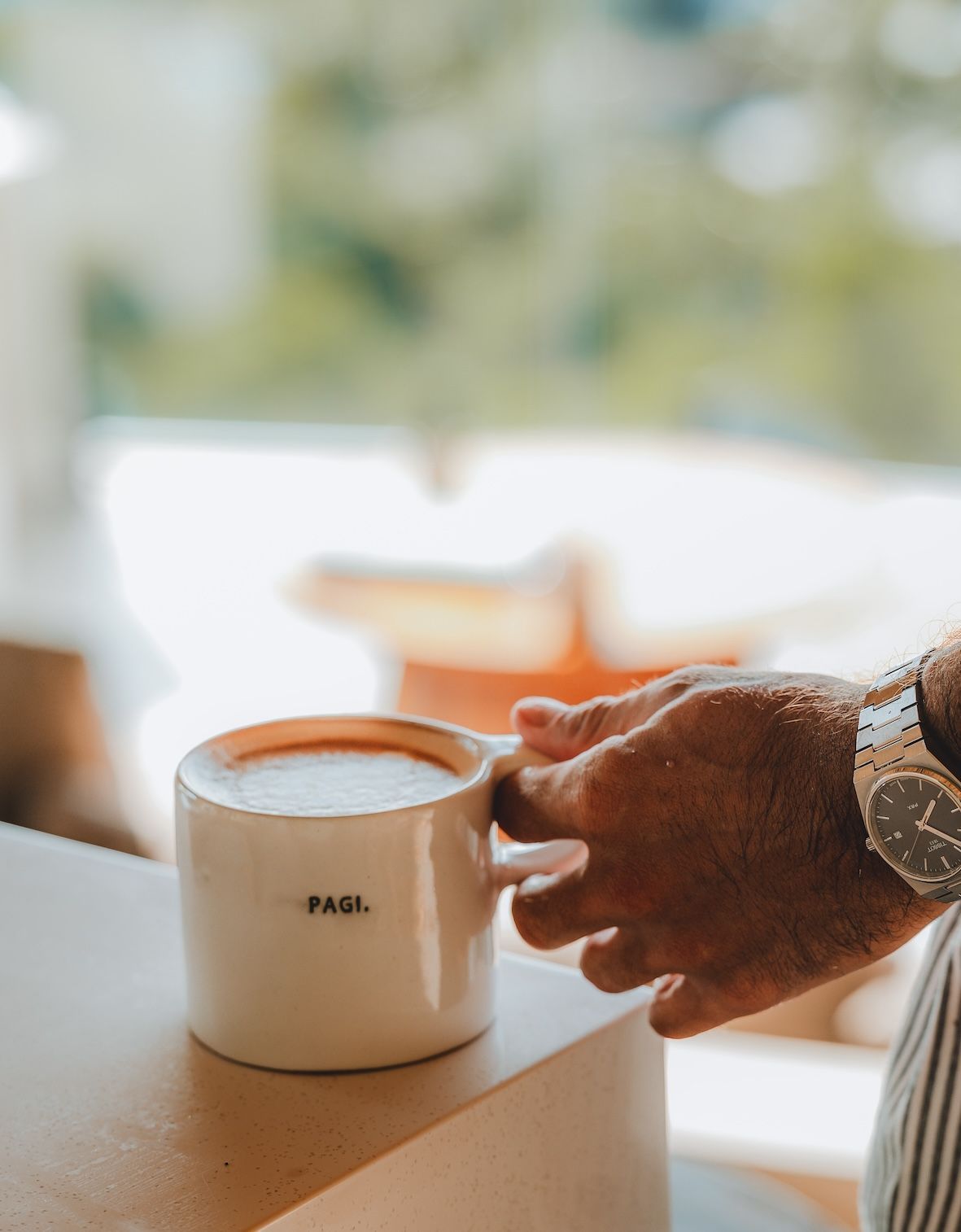 Hand holding a white mug of coffee on a table in a bright, blurred room