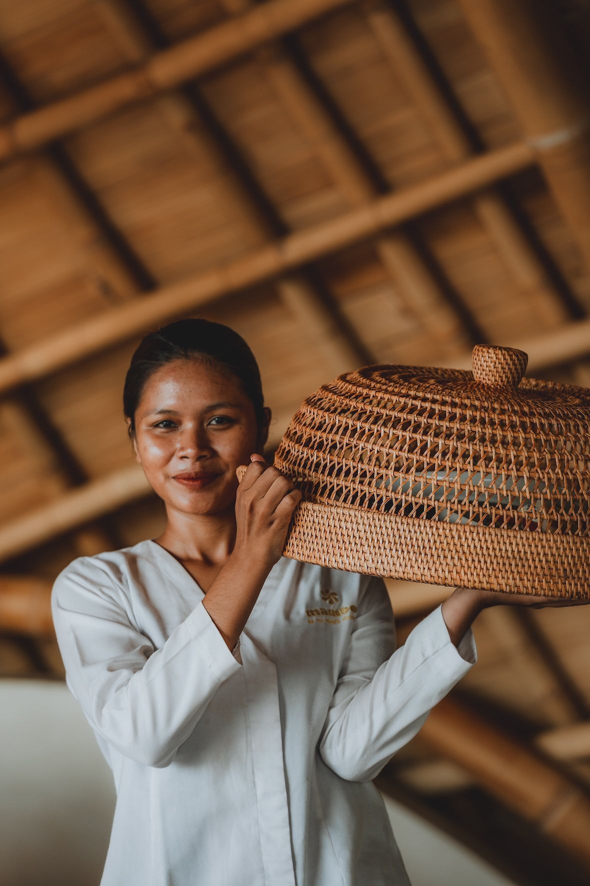 Woman in a white shirt holding a woven basket indoors under a thatched roof