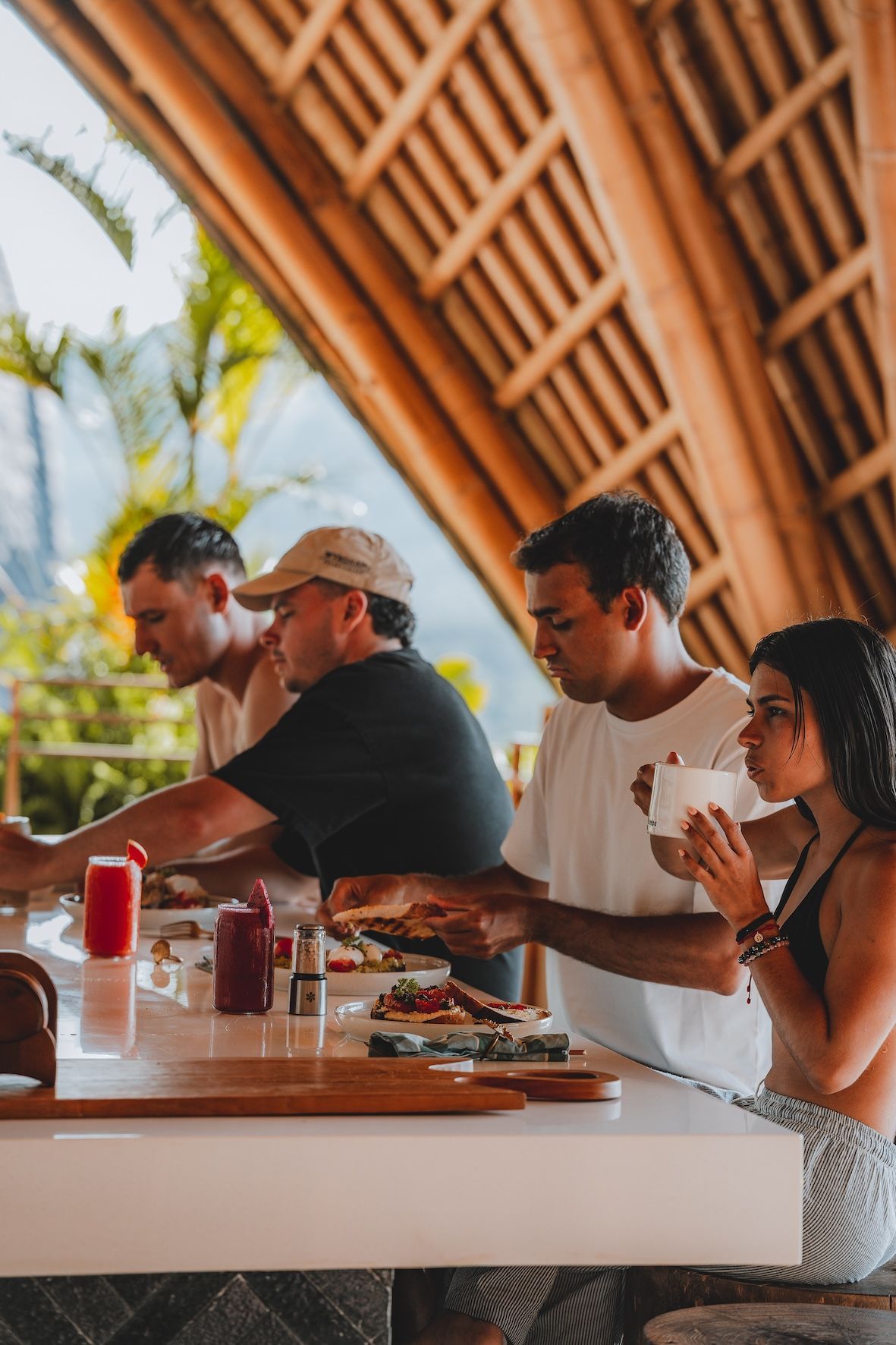 People seated at a wooden bar under a bamboo roof, eating and talking in a tropical setting.