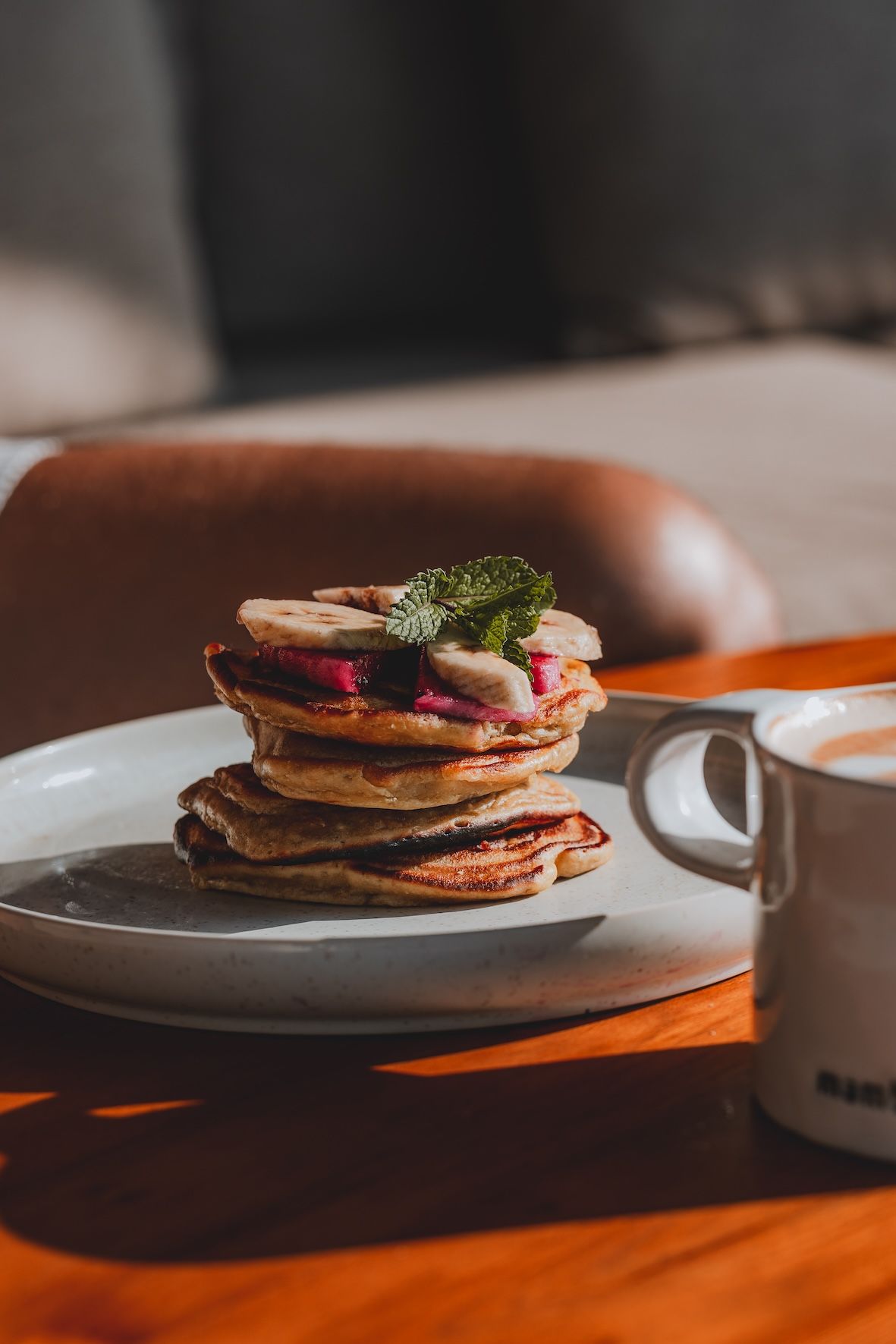 Stack of pancakes with berries and herbs on a white plate beside a coffee mug on a table