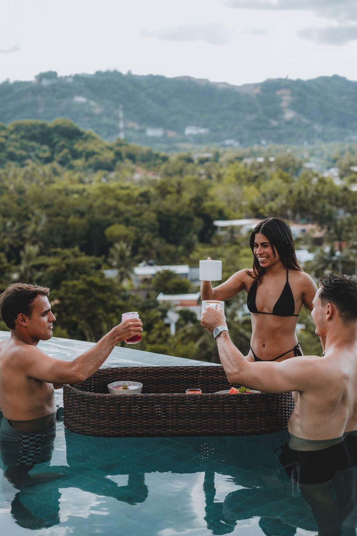 People relaxing in a pool, clinking drinks beside a wicker table with a mountain view.