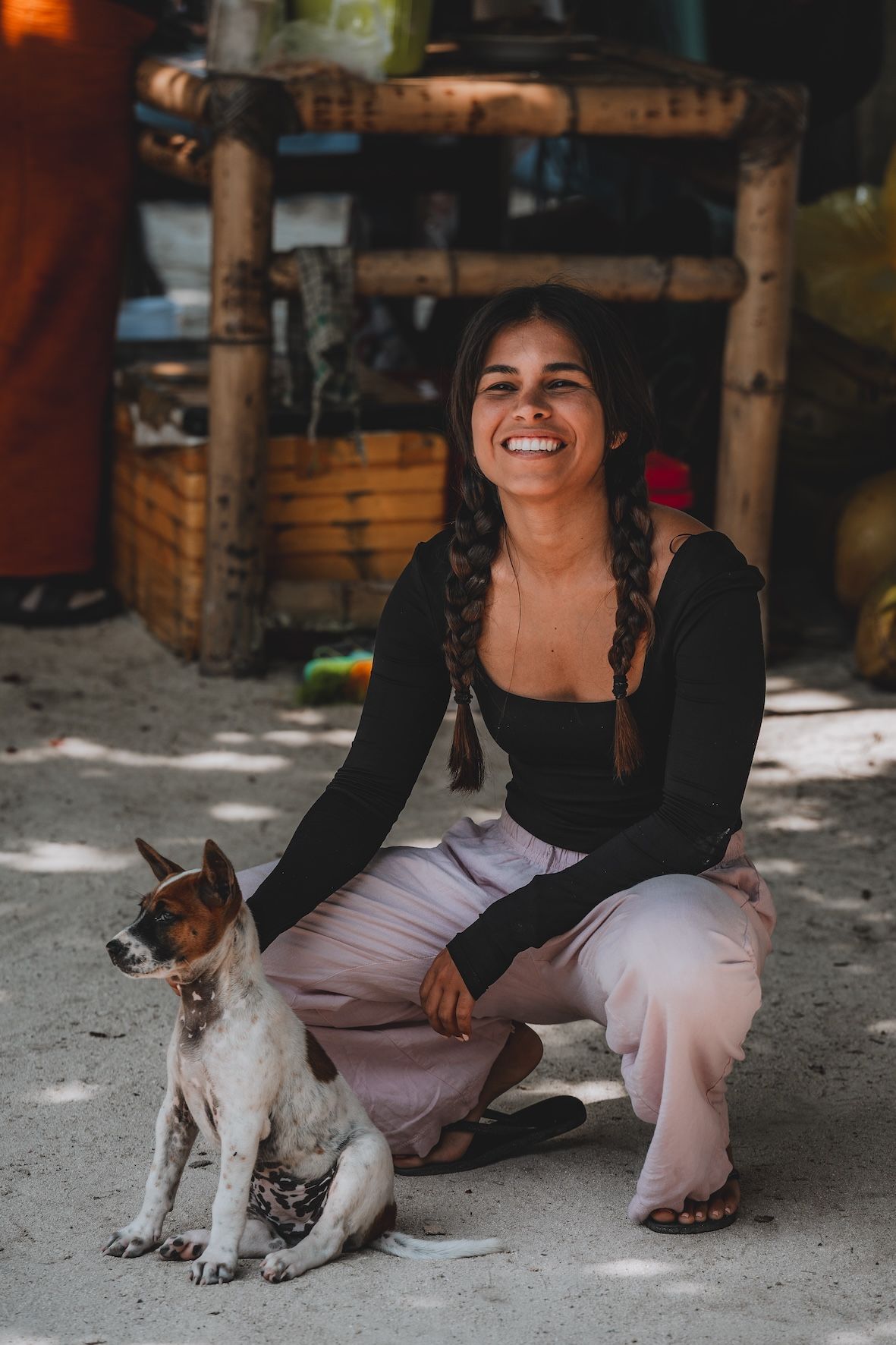 Smiling woman crouches beside a small dog outdoors in a sandy yard