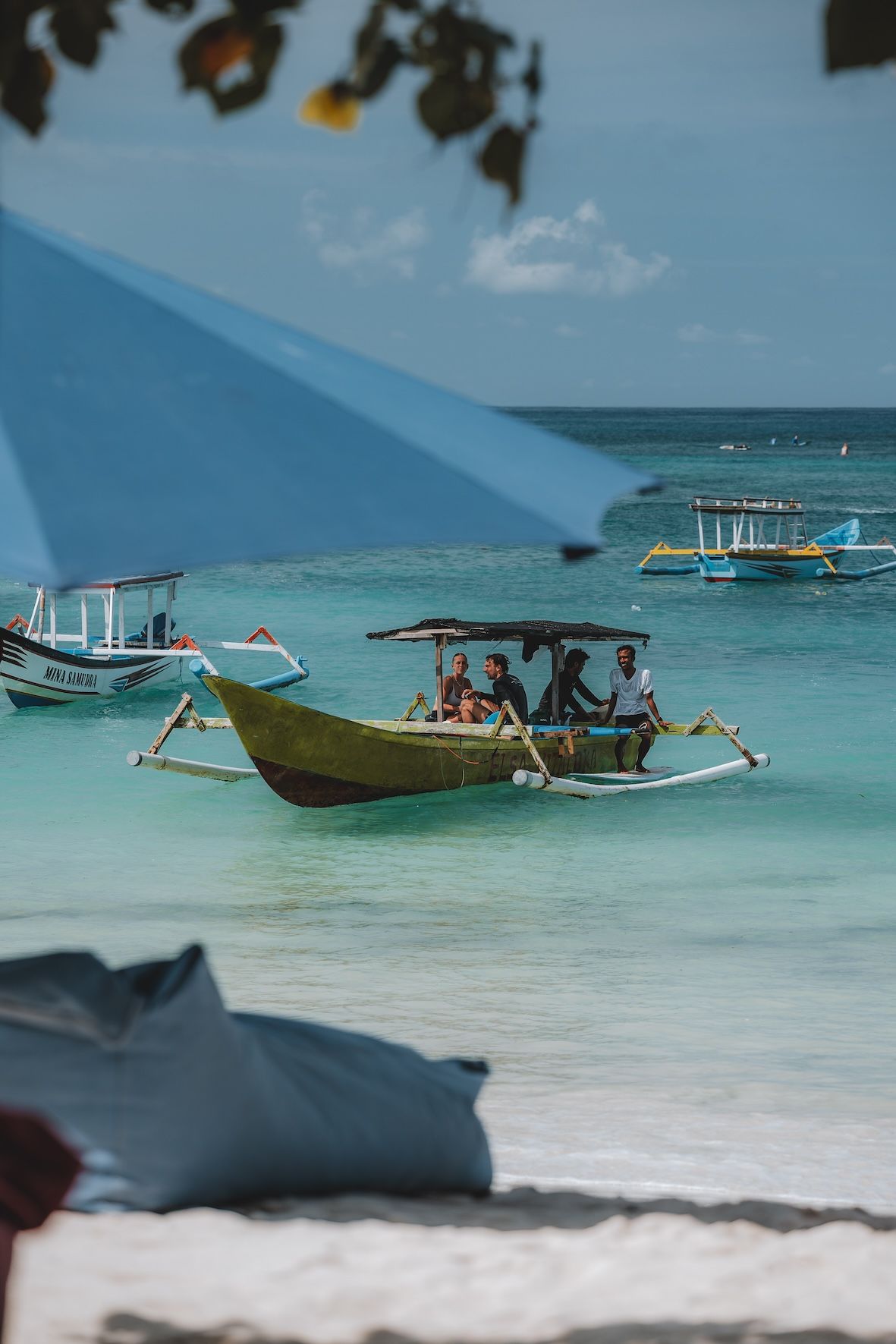 Small boats on clear turquoise water near a beach, with blue sunshade in the foreground.