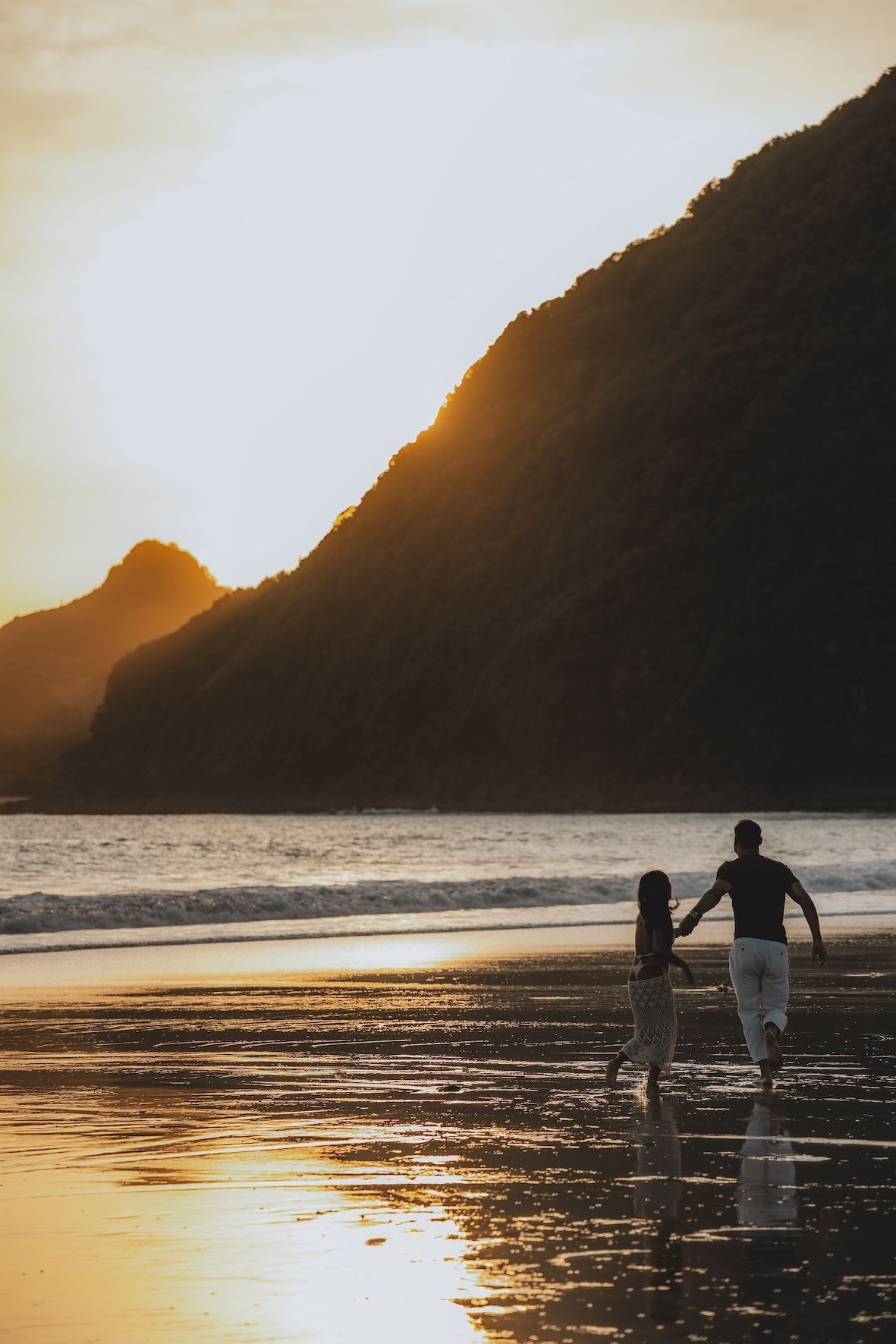 Person walking with a dog on a beach at sunset, with a large dark hill in the background