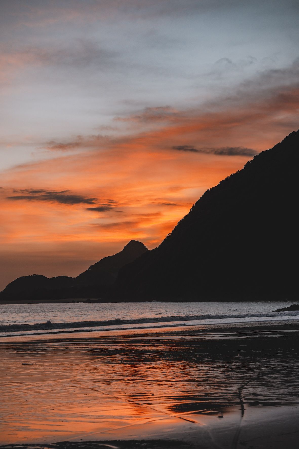 Orange sunset over a calm beach with dark mountains silhouetted against a cloudy sky