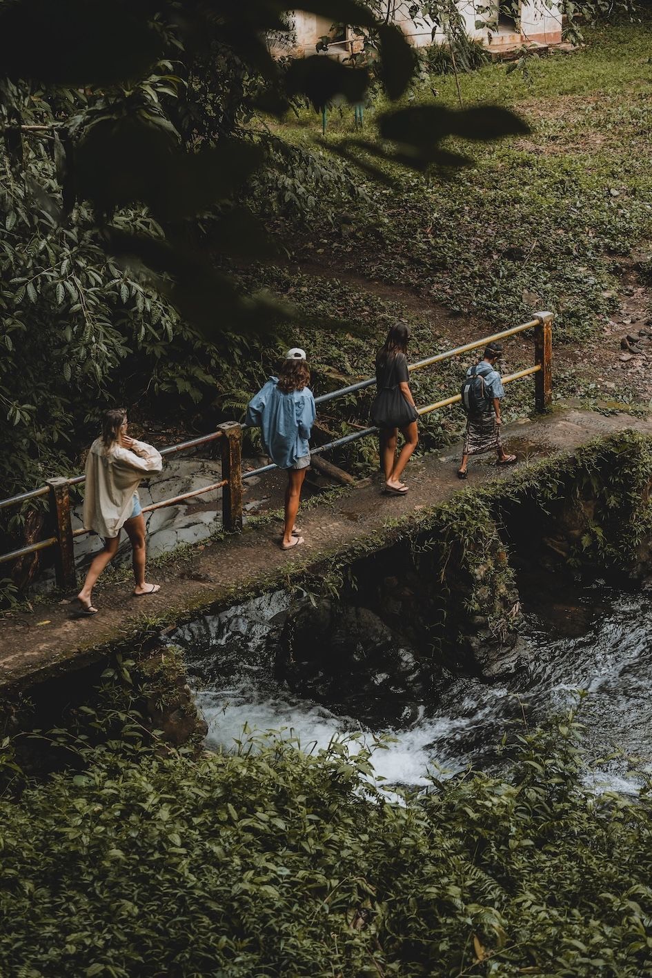 Four people crossing a narrow footbridge over a small creek in a green, wooded area