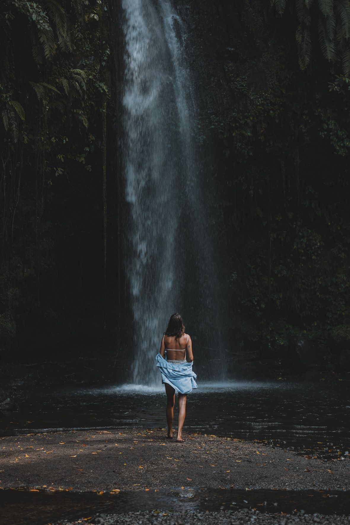 Person standing in a shallow pool before a tall waterfall in a dark forest
