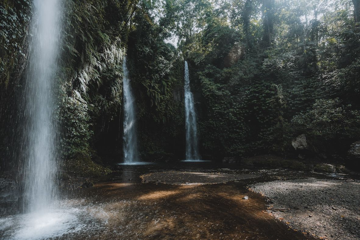Forest waterfall with three slender cascades flowing into a rocky pool, surrounded by dense green trees