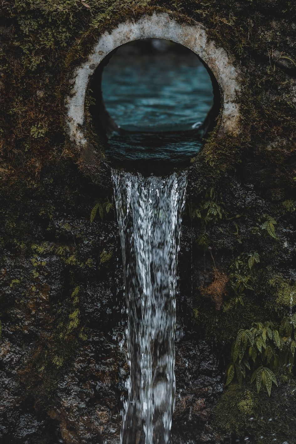 Old stone drain with water pouring into a dark, moss-covered wall opening