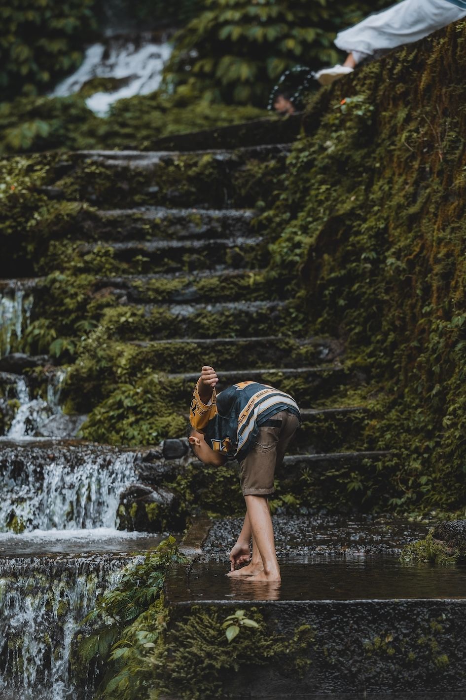Person bending by a small waterfall on moss-covered stone steps in a lush forest setting