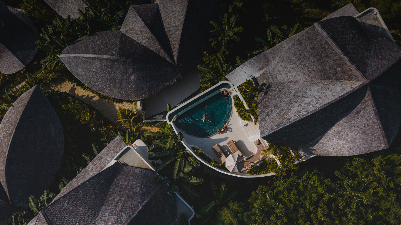 Aerial view of a resort pool surrounded by dark thatched-roof villas and tropical greenery.