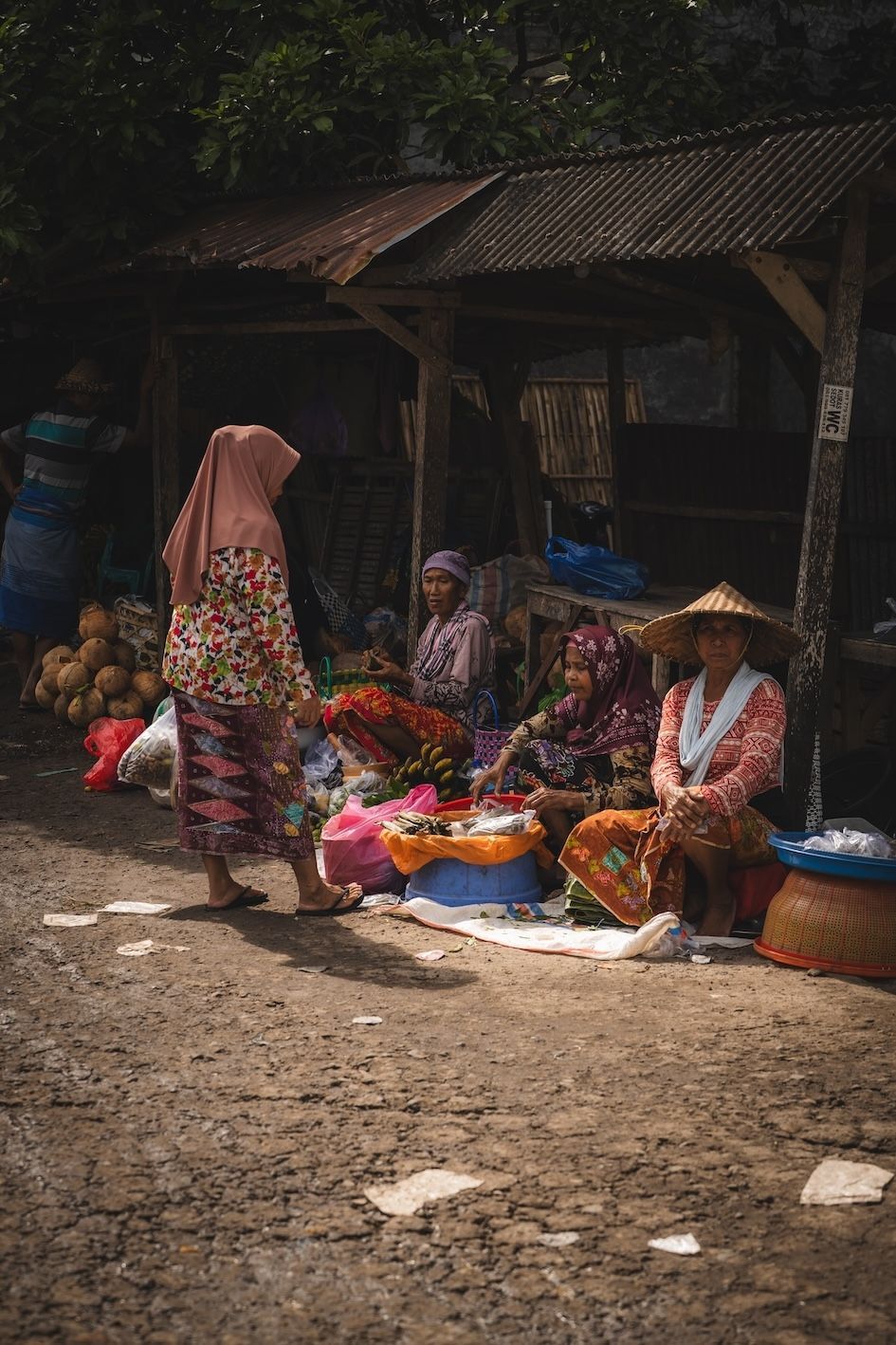 Street market vendors sit among colorful goods outside a rustic shop, while a woman stands nearby on a dirt road.