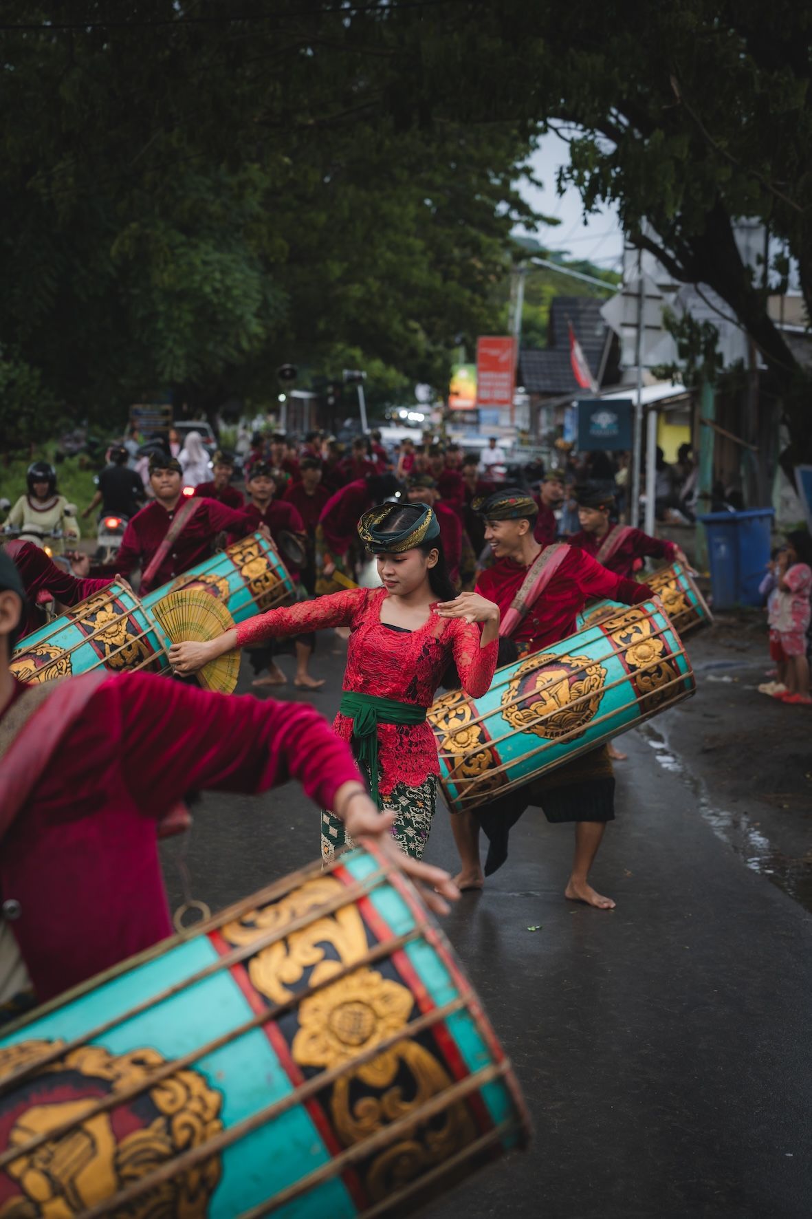 Drummers in red costumes perform a street parade on a wet road under trees