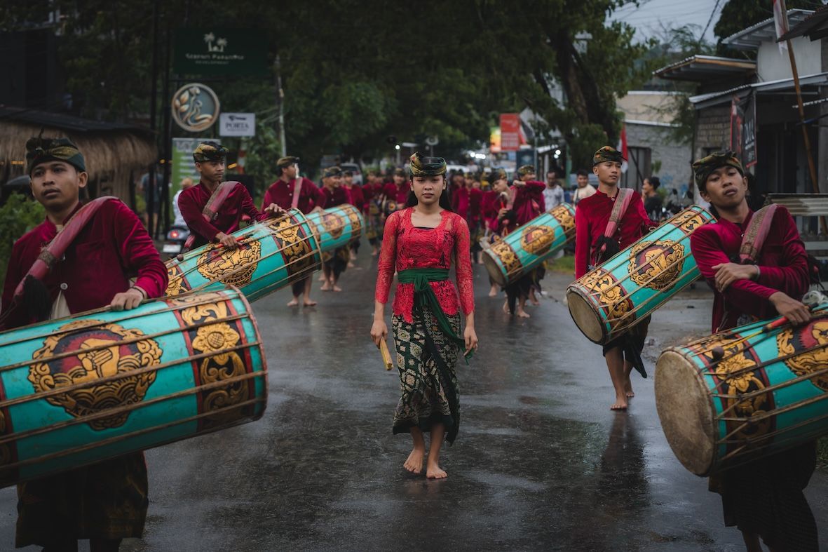 Drummers in red outfits play large turquoise drums while marching down a wet street in a procession