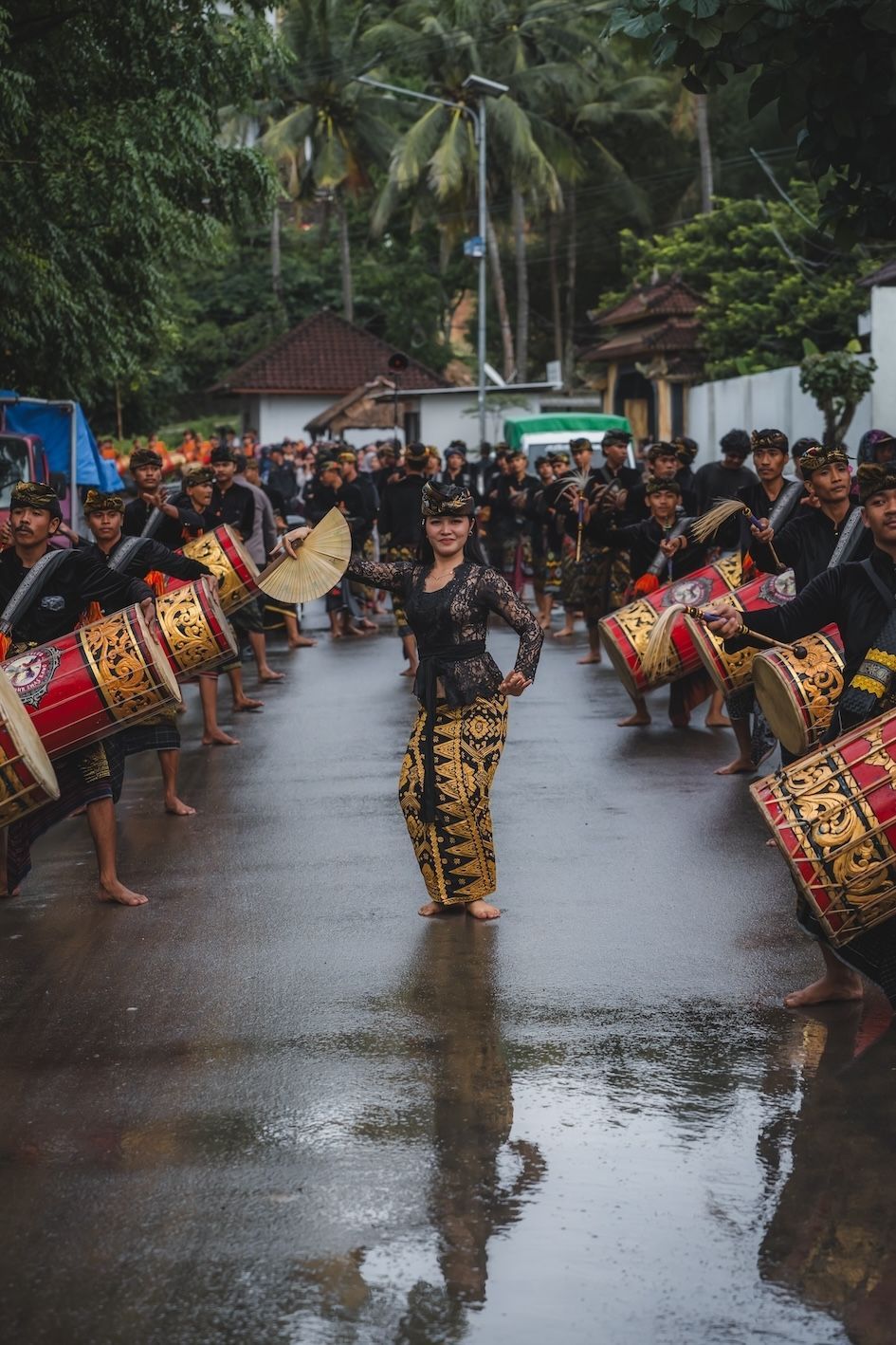 Traditional dancers and drummers perform on a wet road in a tropical village procession