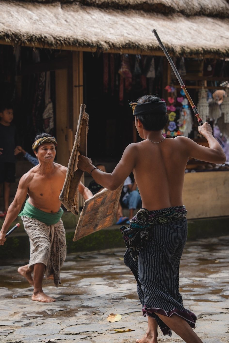 Two men dancing with woven shields and sticks in a muddy village courtyard before thatched huts