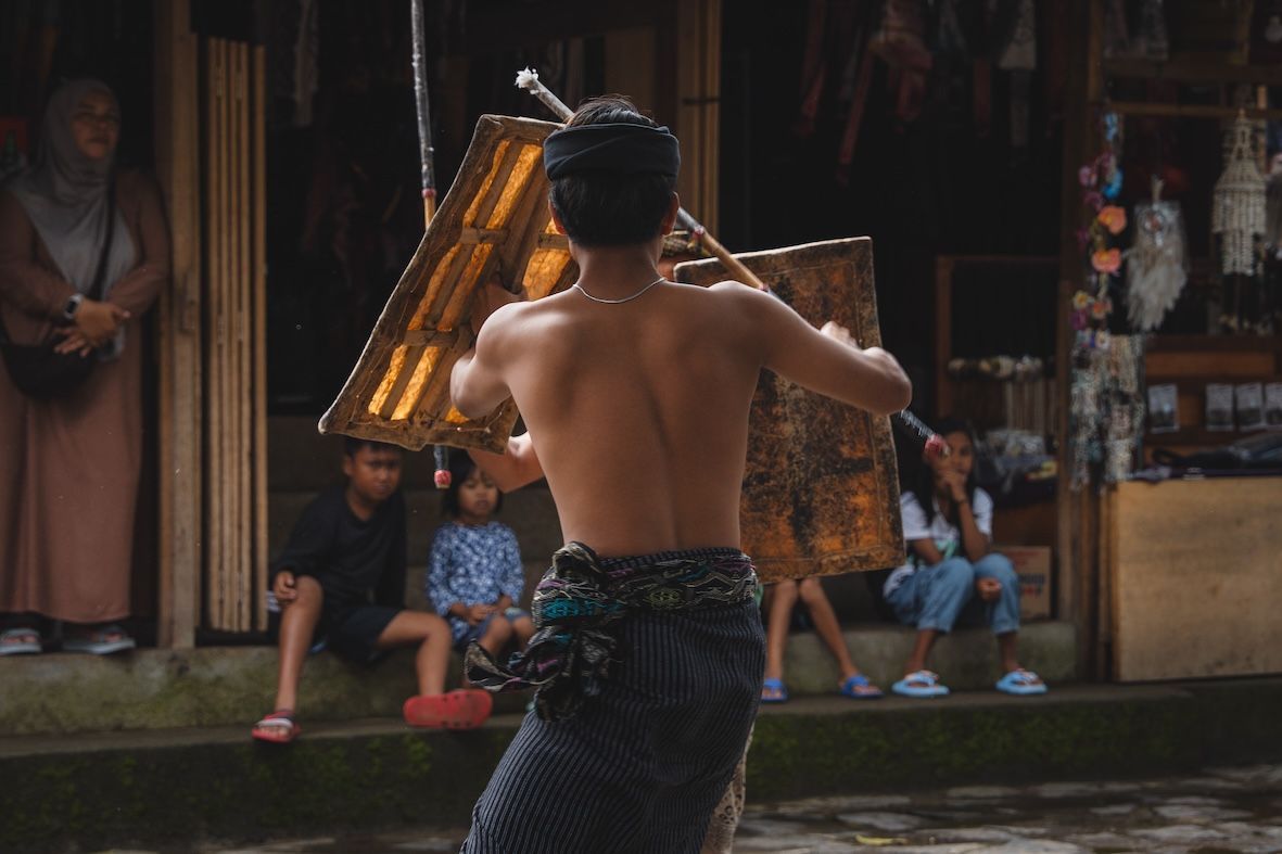 Shirtless performer in a black patterned sarong dances before seated spectators in a dim indoor venue