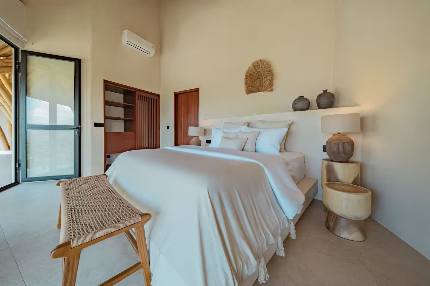 Bright minimalist bedroom with white bedding, woven chair, shelves, and an open door to a balcony