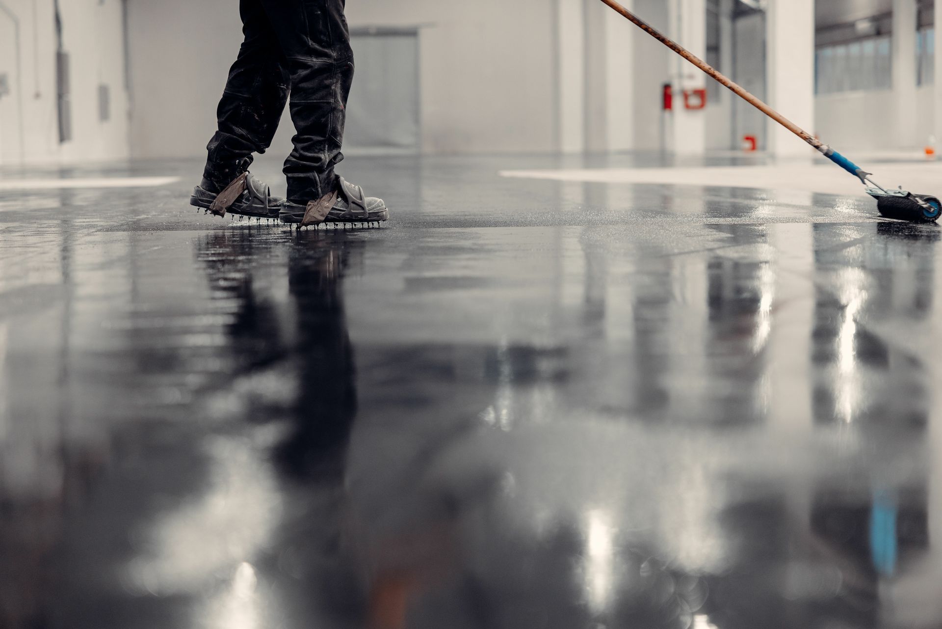 Person applying epoxy coating to a large, glossy floor with a long-handled applicator in a bright, industrial setting. Person applying epoxy coating to a large, glossy floor with a long-handled applicator in a bright, industrial setting.
