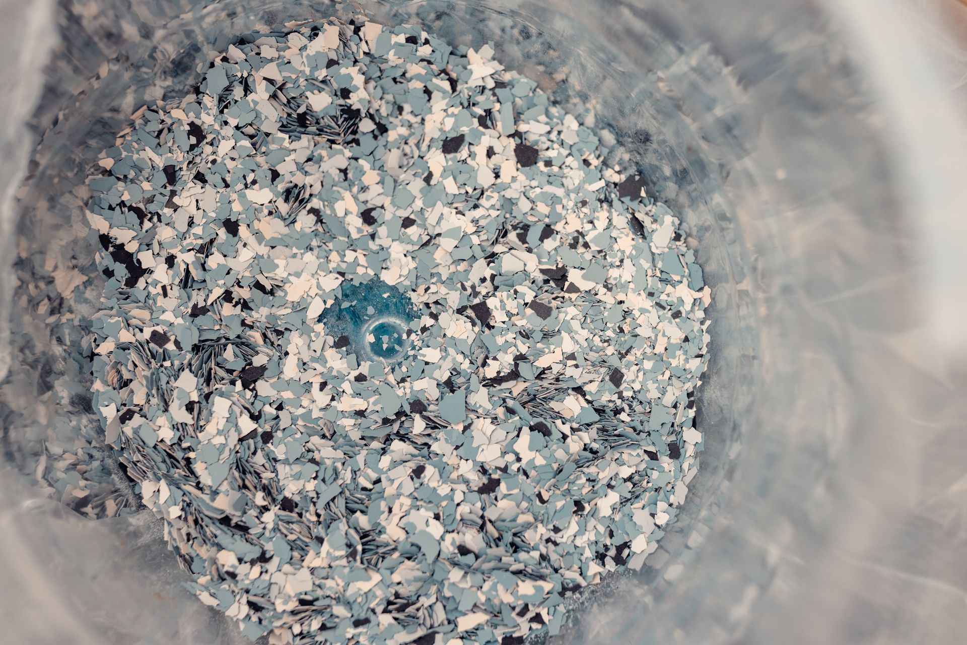 Blue, white, and black floor epoxy flakes in a clear plastic container, seen from above. Blue, white, and black floor epoxy flakes in a clear plastic container, seen from above.