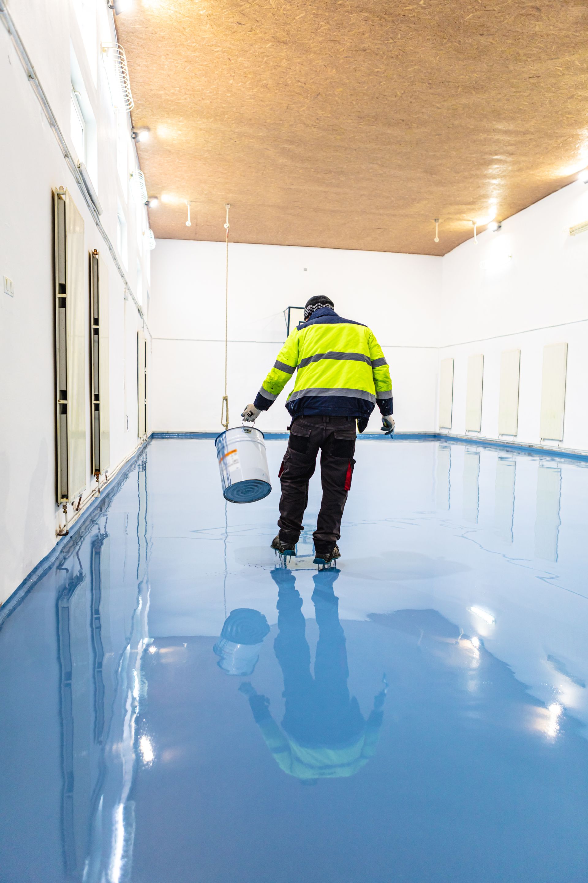 Person in a reflective jacket walks on a blue epoxy floor, holding a paint can. Long white room, high ceiling.