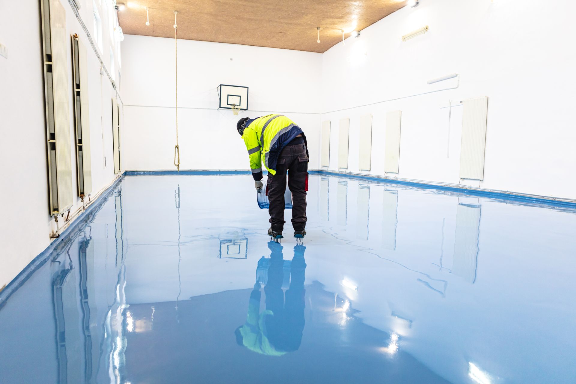 Worker applying blue epoxy to a spacious room's floor. Reflective surface, white walls, and a basketball hoop.