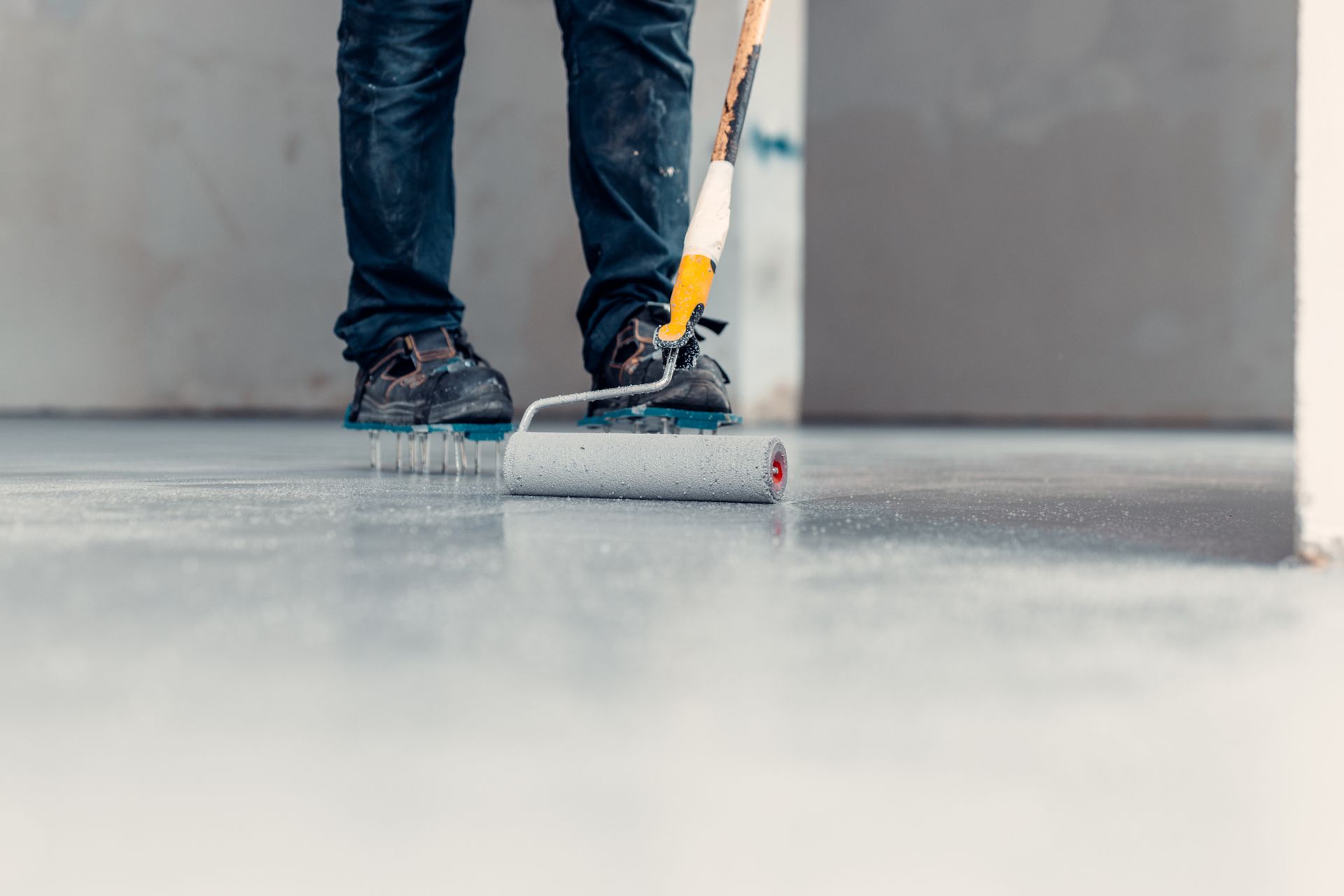 Person in jeans and work boots rolling grey paint onto a concrete floor. Person in jeans and work boots rolling grey paint onto a concrete floor.