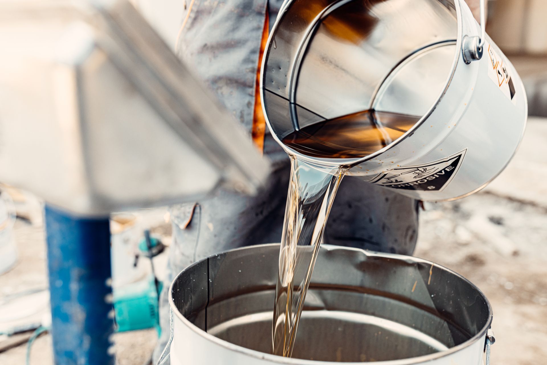 Worker pouring liquid from a metal bucket into a larger metal container. Worker pouring liquid from a metal bucket into a larger metal container.