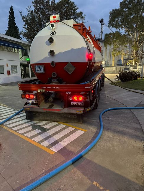 Un camión cisterna rojo y blanco estacionado en un terreno pavimentado al anochecer, con mangueras en el suelo.