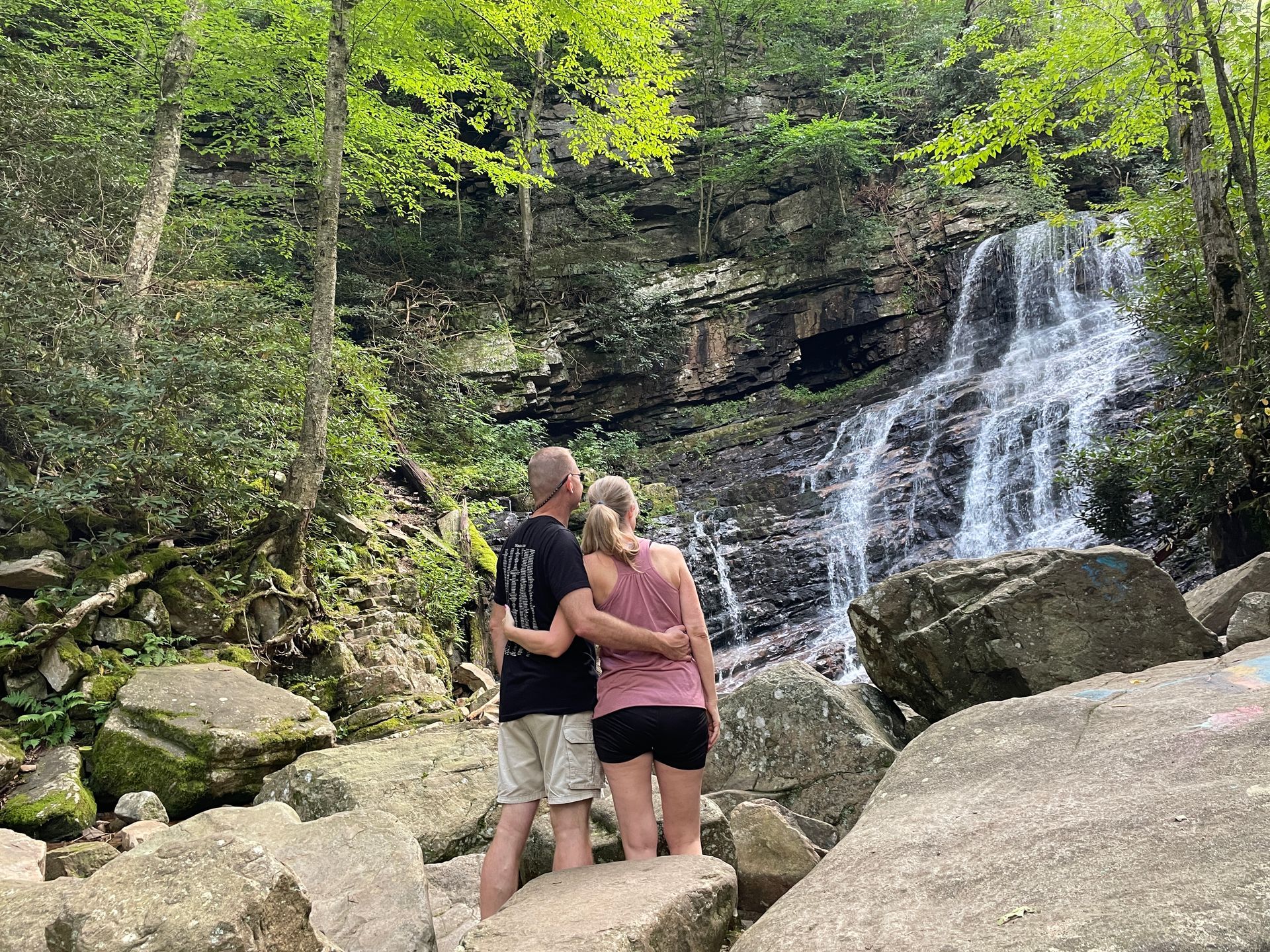 Couple embraces, looking at a waterfall. They stand on rocks amid lush trees.