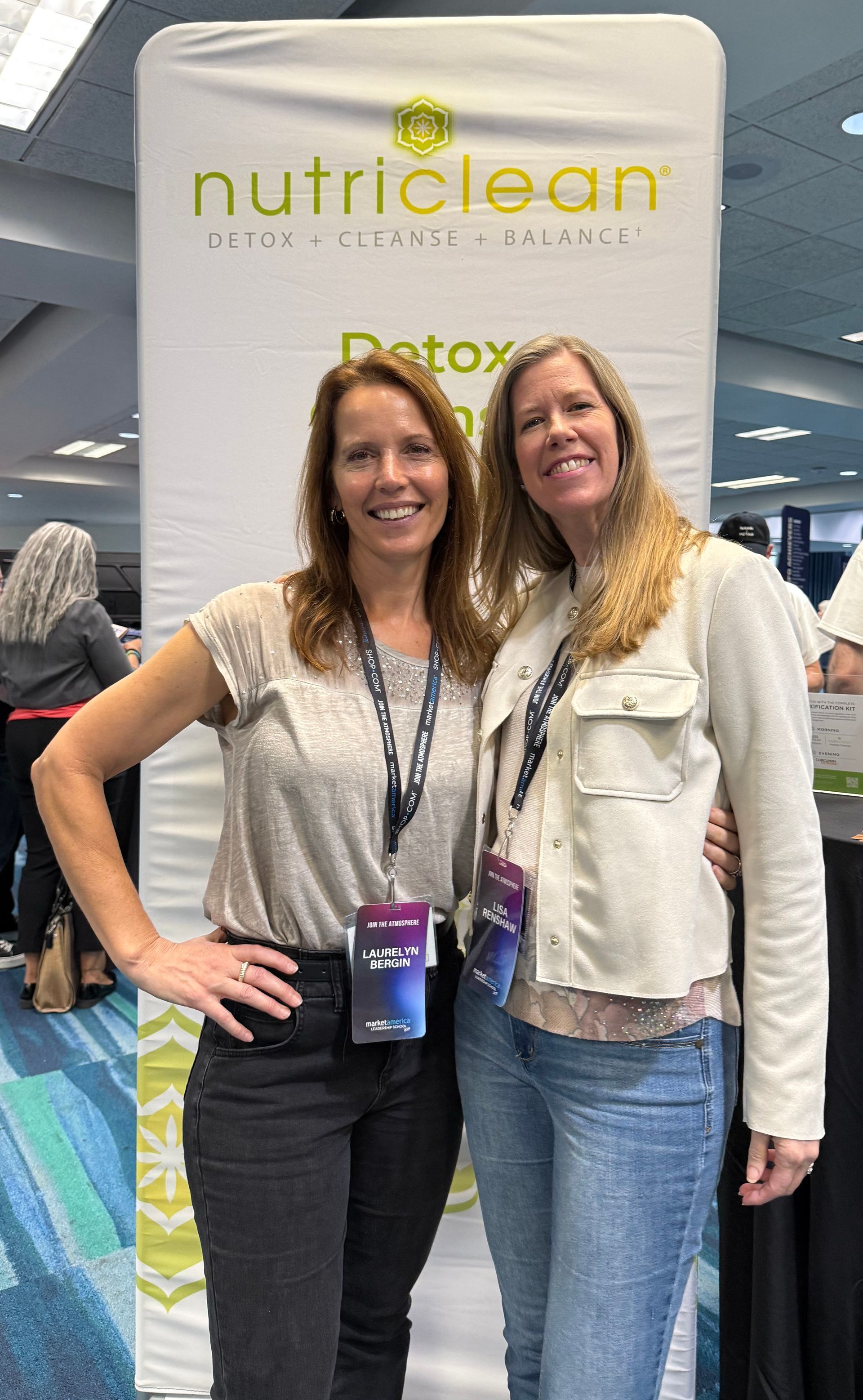 Two women smiling, posing in front of a “NutriClean” banner at an event.