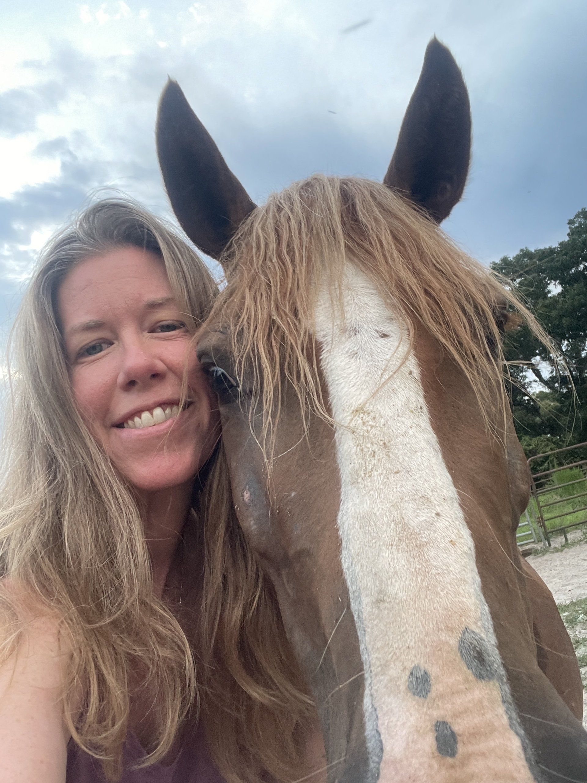 Woman smiling next to a brown and white horse with wet mane; outdoors under cloudy sky.