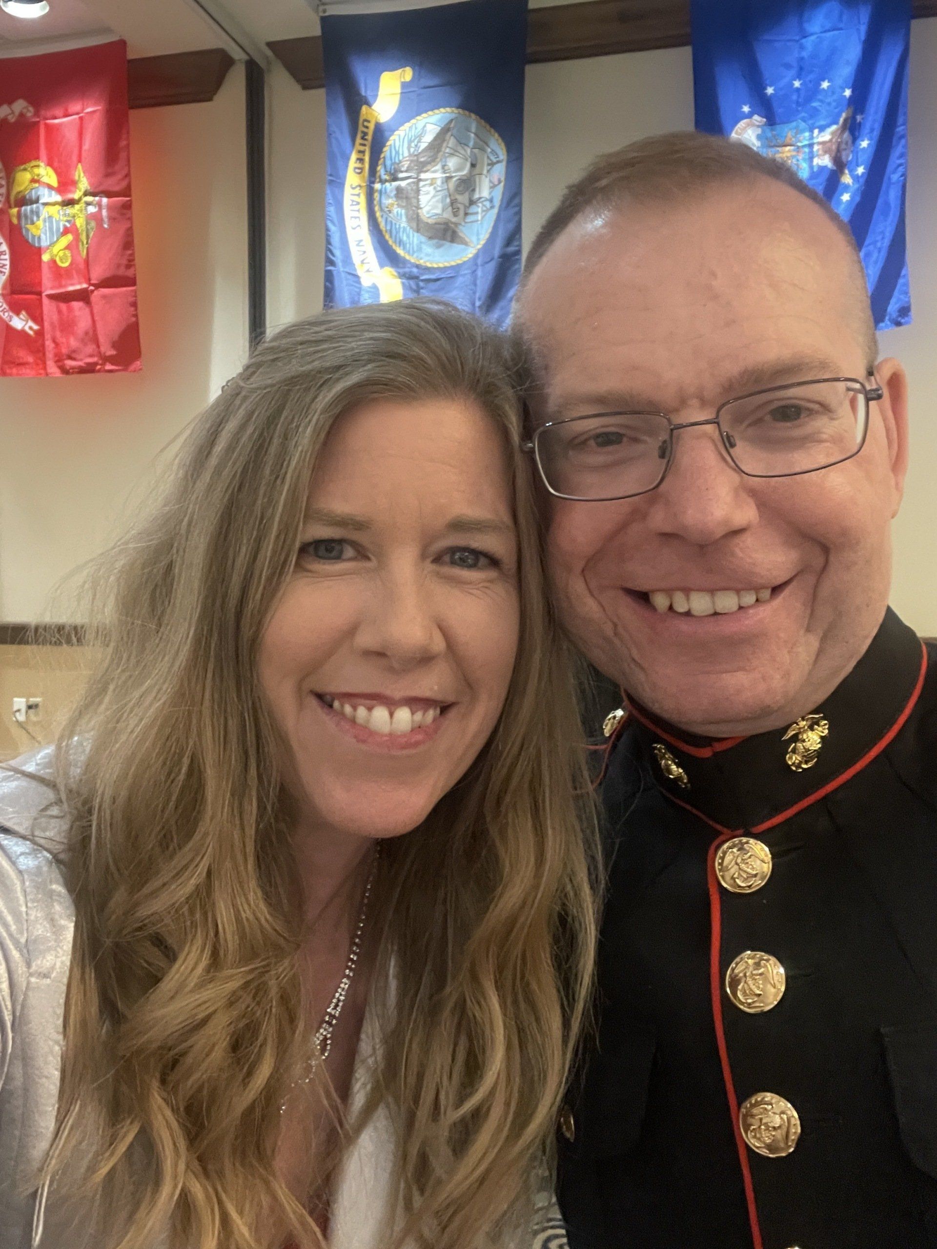 Woman and man in uniform smiling at the camera, Marine Corps jacket, flags in the background.