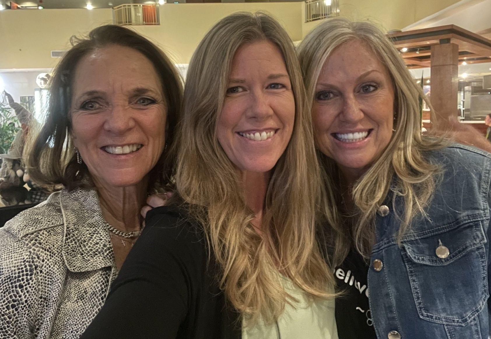 Three women smiling, posing for a photo indoors, one wearing a denim jacket.