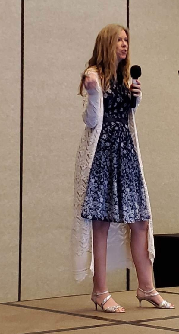 Woman in a floral dress and lace cardigan speaks into a microphone on a stage.