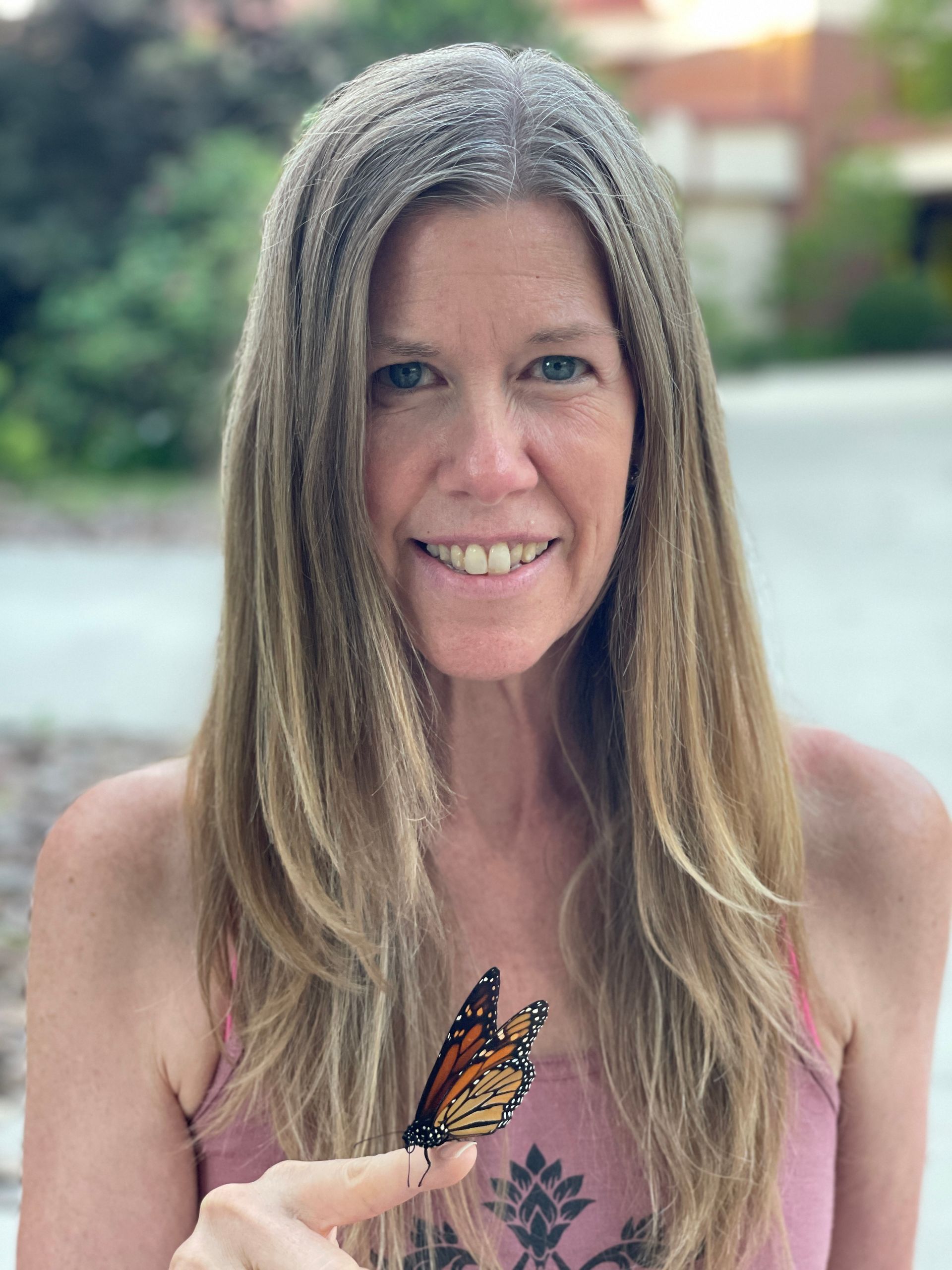 Woman with long blonde hair holding a monarch butterfly on her finger, smiling outside.