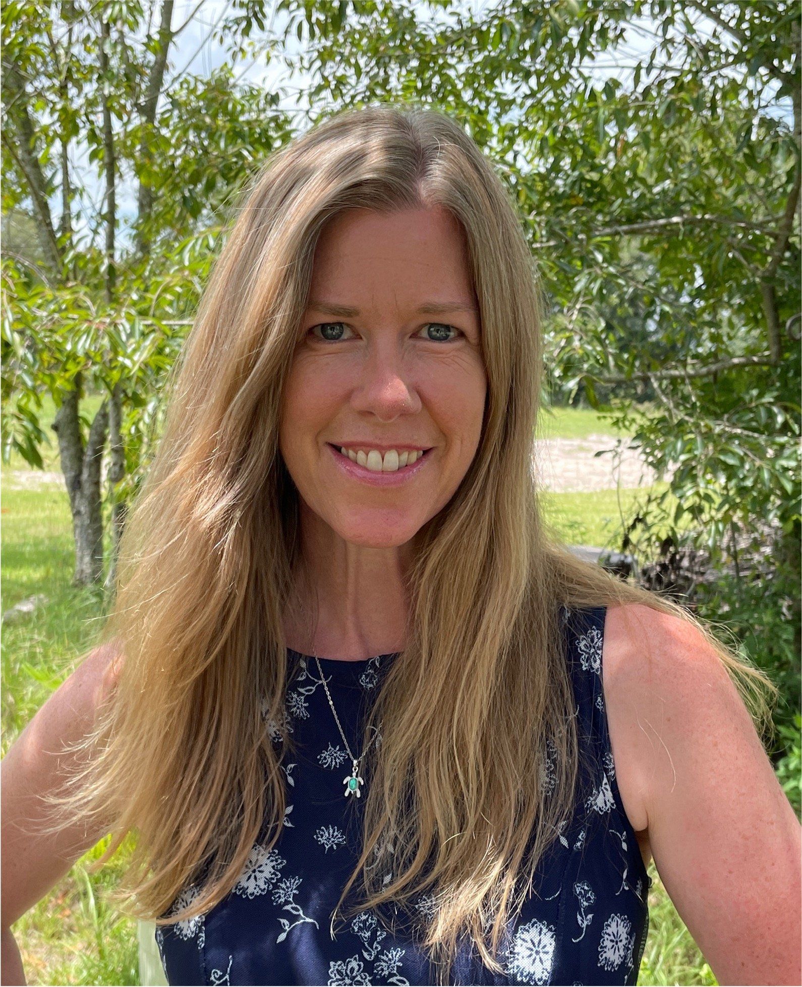 Woman with long blonde hair smiles outdoors, wearing a dark floral top, greenery in background.
