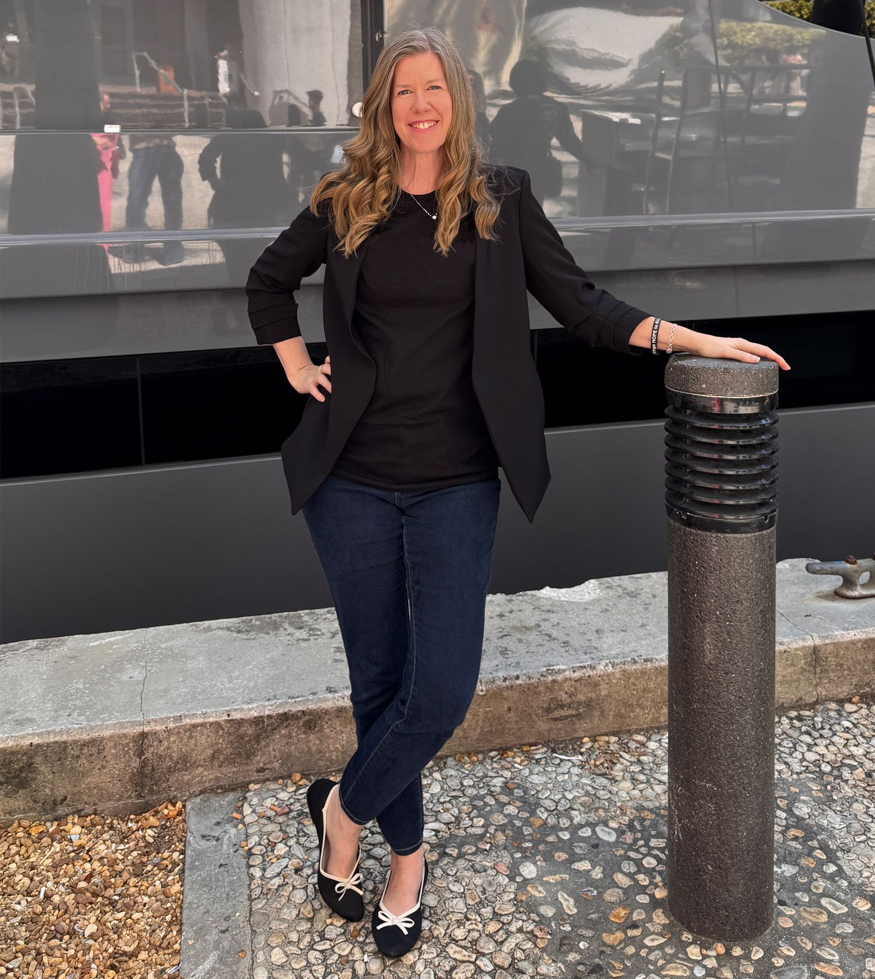 Woman in black blazer, jeans, and flats leaning on a pillar, smiling. Outdoors, by water.