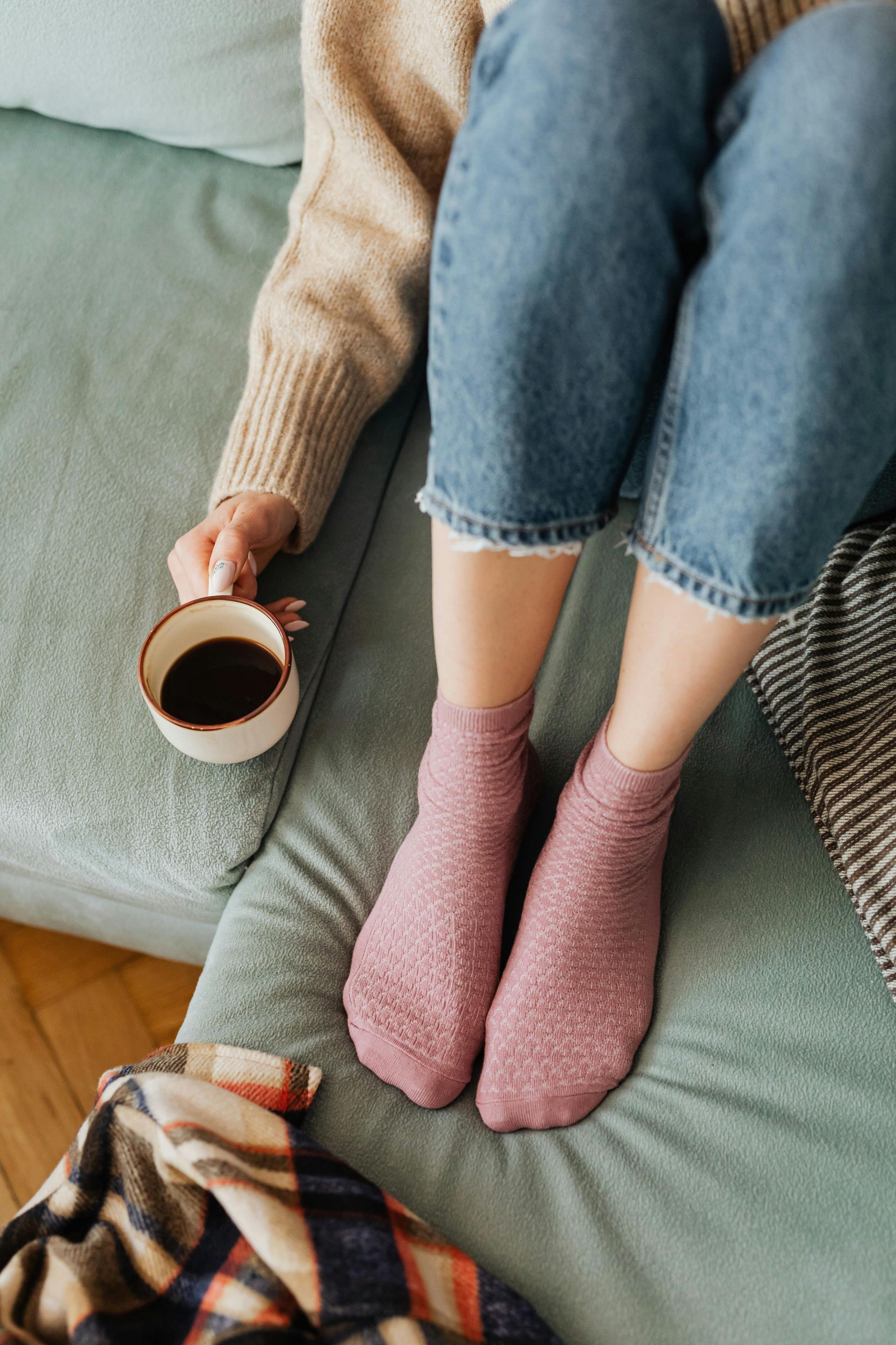 Una persona seduta su un divano color verde acqua, con indosso calzini rosa pelosi e jeans, tiene in mano una tazza di caffè, con una coperta a fantasia nelle vicinanze.