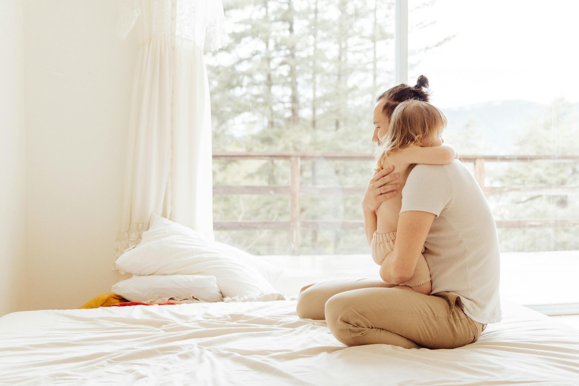 Una persona è inginocchiata su un letto bianco, abbracciando dolcemente un bambino piccolo, immerso nella luce soffusa proveniente da una grande finestra vicina.