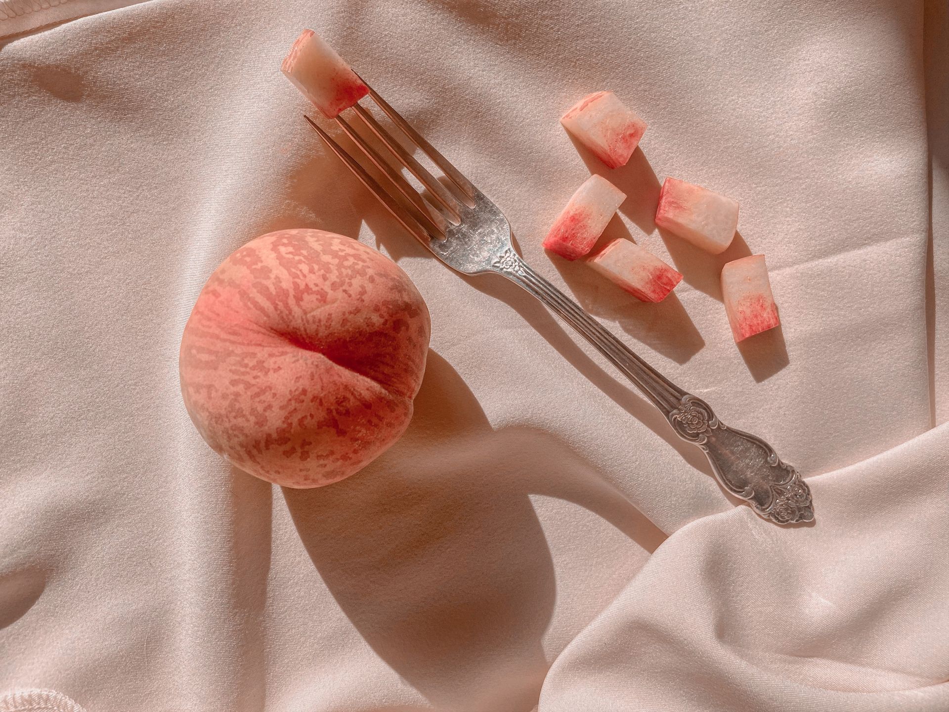Una pesca intera e diversi pezzi tagliati sono adagiati su un morbido tessuto rosa pallido accanto a una forchetta di metallo decorata.
