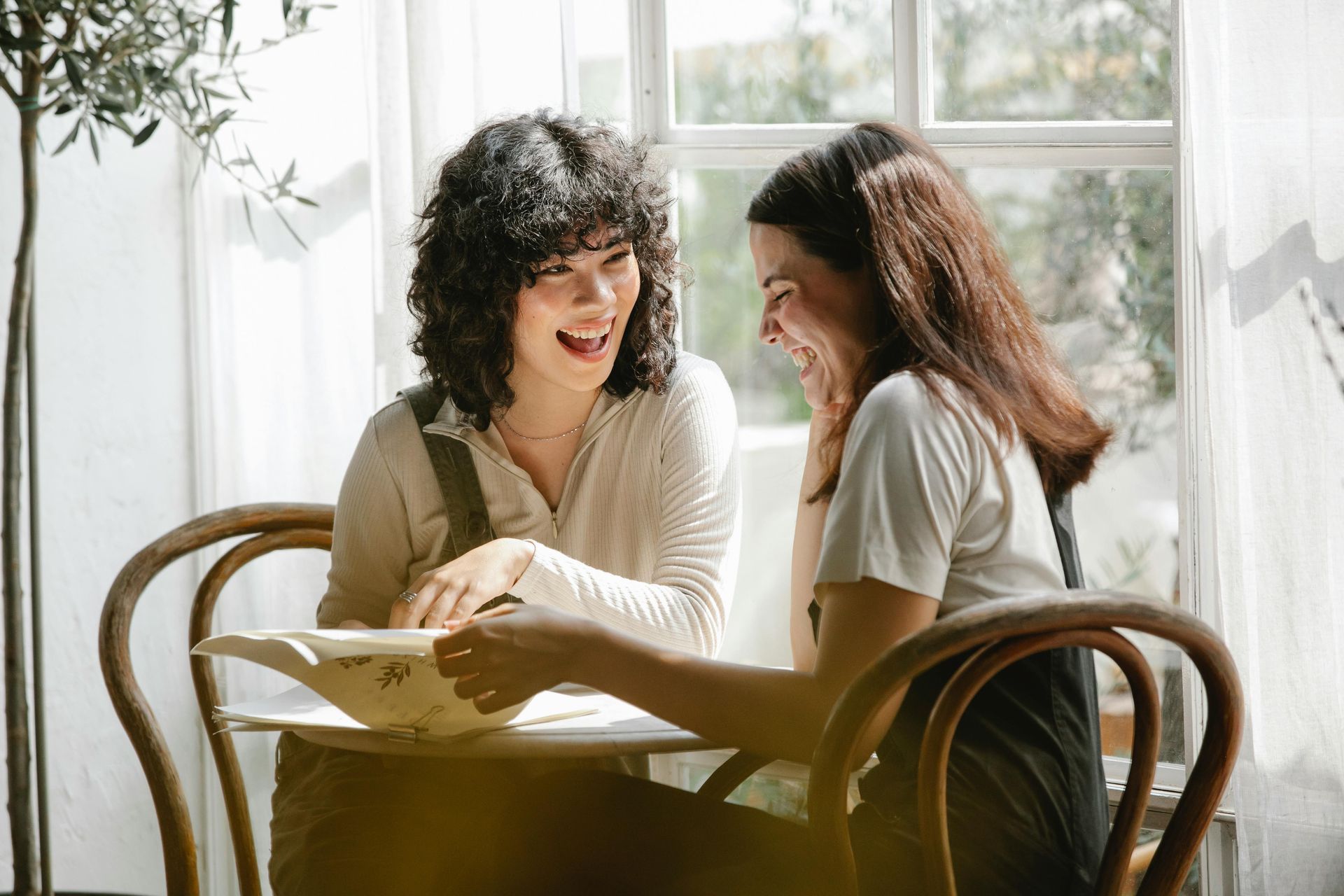 Due persone siedono a un tavolino vicino a una finestra luminosa, sorridono e guardano insieme un libro aperto.