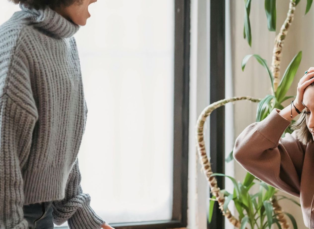 In un ambiente interno e luminoso, una persona con un maglione grigio urla contro un'altra che appare stressata, con le mani tra i capelli.